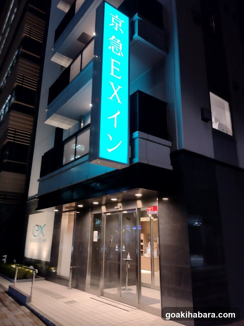 Night view of the exterior entrance of a modern building with illuminated vertical signage reading '京急E Xイン' (Keikyu E-X Inn). The facade features glass doors, dark cladding, and balconies. A smaller logo is visible on the left wall near ground level.