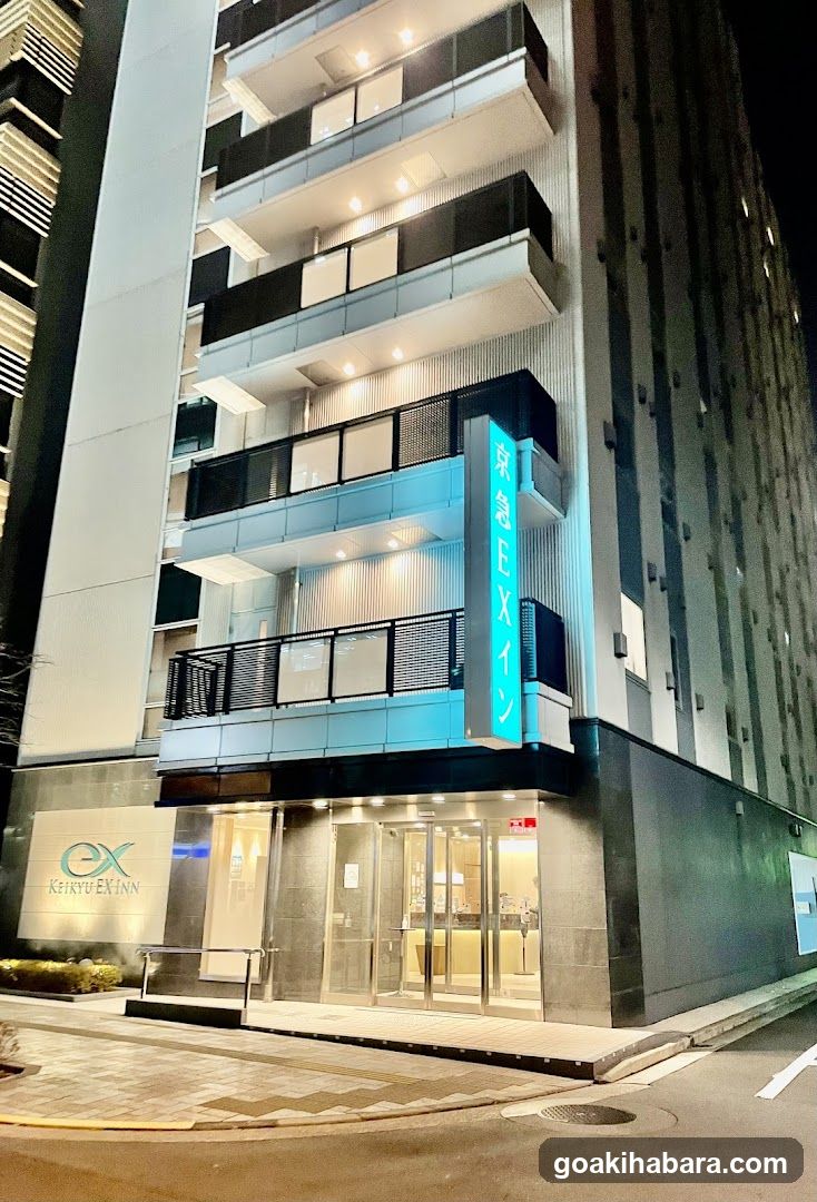 Night view of the exterior entrance and storefront facade of a modern multi-story building with illuminated signage reading 'Kinkyu Ekinn' on the side and logo at ground level. Glass doors lead into lobby area.