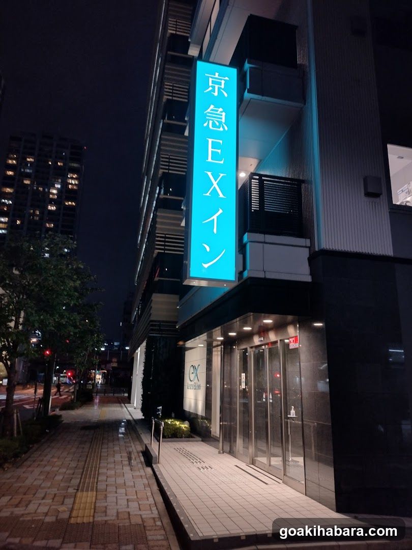 Night view of a modern building entrance with illuminated vertical signage reading '京急EXイン' (Kyoto Express Inn). The storefront features glass doors and windows, with visible interior lighting. A paved sidewalk runs alongside the building, bordered by trees and streetlights in the background.