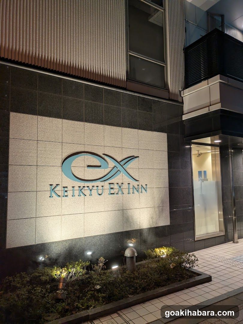 Night view of the exterior facade featuring a large illuminated sign with 'ex' logo and 'KEIKYU EX INN' text on tiled wall, flanked by modern building architecture and small landscaped garden area.