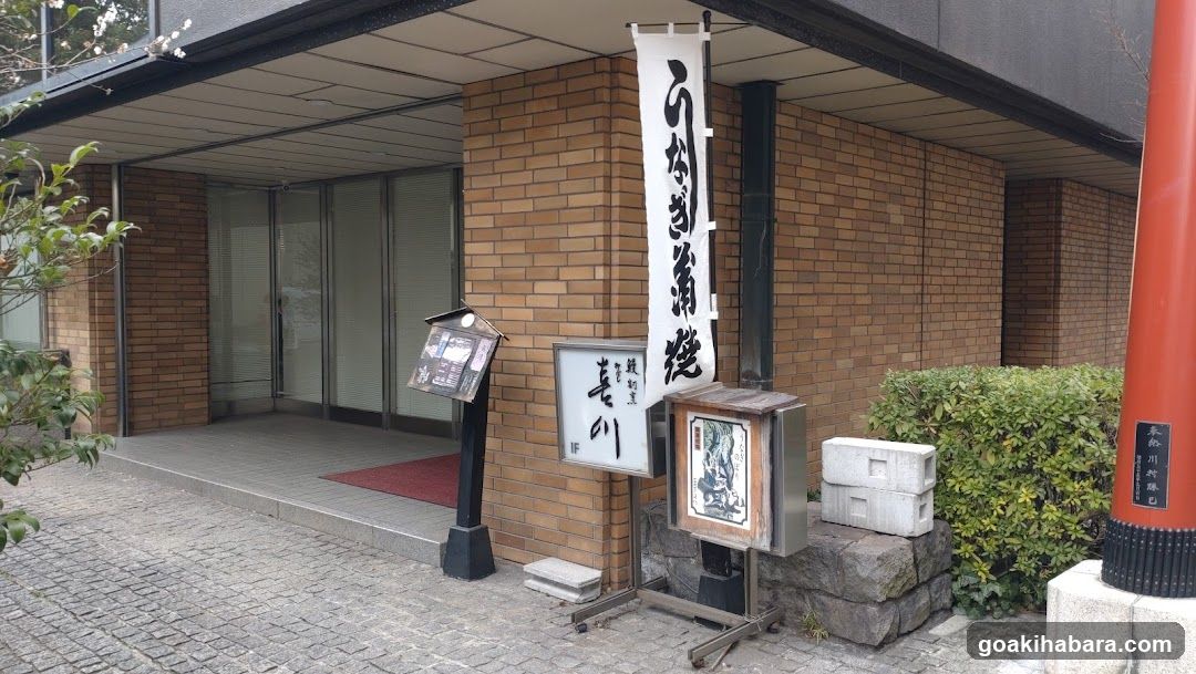 The image shows the exterior entrance of a building with brick walls and glass doors. There is a vertical banner with Japanese calligraphy hanging on the wall, and a smaller signboard displaying additional text. A menu stand is positioned near the entrance, along with some stacked boxes and greenery nearby.