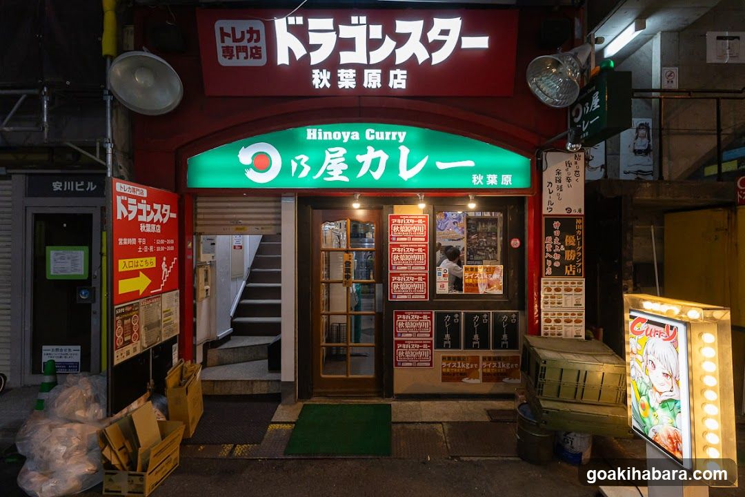 Night shot of the storefront featuring a red sign with 'Dragon Star' and green neon signage reading 'Hinoya Curry'. The entrance has glass doors, posters on the wall, and stacked boxes outside.