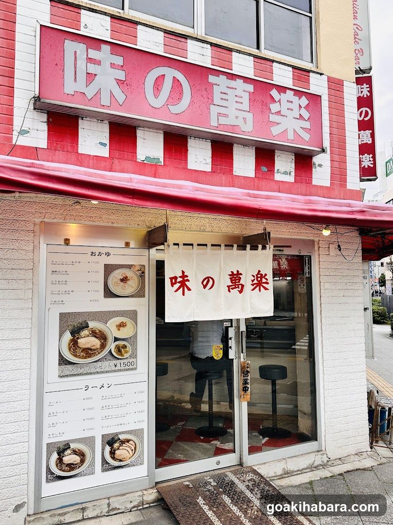 A shop front with a red and white striped facade, featuring large Japanese characters on the sign above the entrance. A menu board is visible to the left of the door, displaying various ramen dishes with prices.