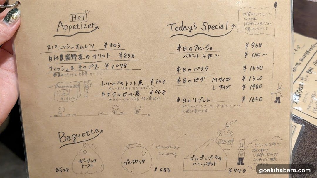 A close-up photo of a hand-drawn menu on brown paper, held by a person with glittery nail polish. The menu includes sections for 'Hot Appetizer', 'Today's Special', and 'Baguette'. Items are written in Japanese with prices in yen.