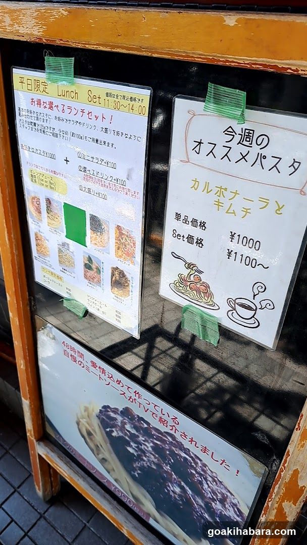 A close-up view of a restaurant's exterior, featuring two prominent menu boards taped to the glass door. The left board lists lunch set options with prices and images of dishes. The right board highlights weekly specials like 'Calzone and Kimchi' with pricing details. Below these are additional promotional posters.