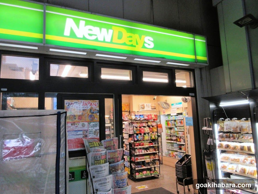 A brightly lit NewDays convenience store entrance with a green and yellow illuminated sign above the door. The storefront features glass doors and windows, displaying various snacks and products inside. A stack of shopping carts is visible near the entrance, along with promotional posters and flyers on display racks outside.