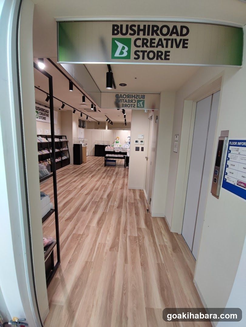 A view looking into a retail store through an open doorway. A large overhead sign reads 'BUSHIROAD CREATIVE STORE' with a green logo. The interior features light wood flooring, track lighting, and shelving units displaying merchandise. A smaller sign is visible further inside, and an elevator is on the right wall.