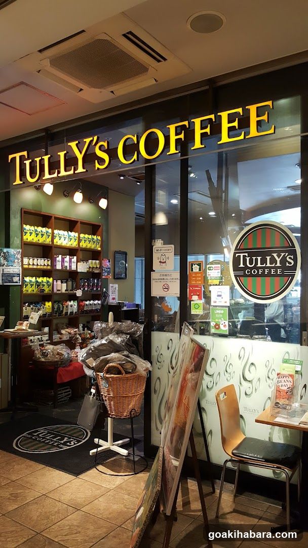 The image shows the exterior entrance of a Tully's Coffee shop. The large yellow 'TULLY'S COFFEE' sign is mounted above the glass facade. Inside, shelves display packaged coffee products and snacks. A wicker basket filled with bread sits near the door. Posters and notices are affixed to the window. A small table with a menu or flyer is visible in the foreground.