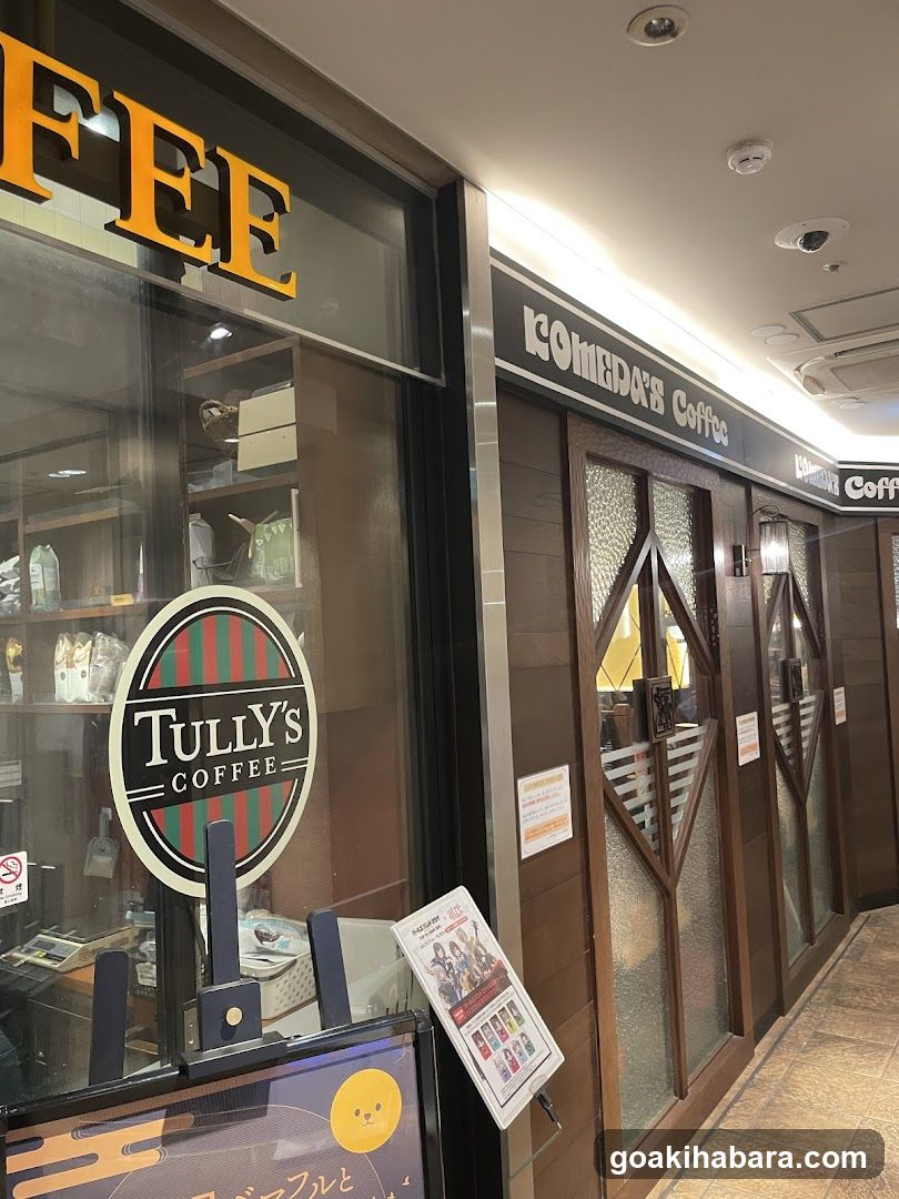 The image shows the exterior of a Tully's Coffee shop, with visible signage reading 'TULLY'S COFFEE' on the window and 'COMEDIA's Coffee' above the entrance. The storefront features glass doors with frosted panels and wooden framing. A promotional standee is placed outside near the entrance.