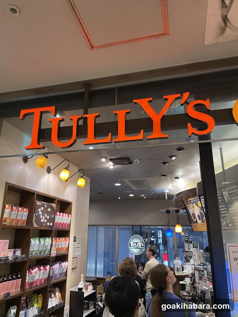 The image shows the exterior of a Tully's coffee shop, with its distinctive orange signage prominently displayed above the entrance. Inside, shelves stocked with packaged products are visible on the left, while customers gather near the counter area.
