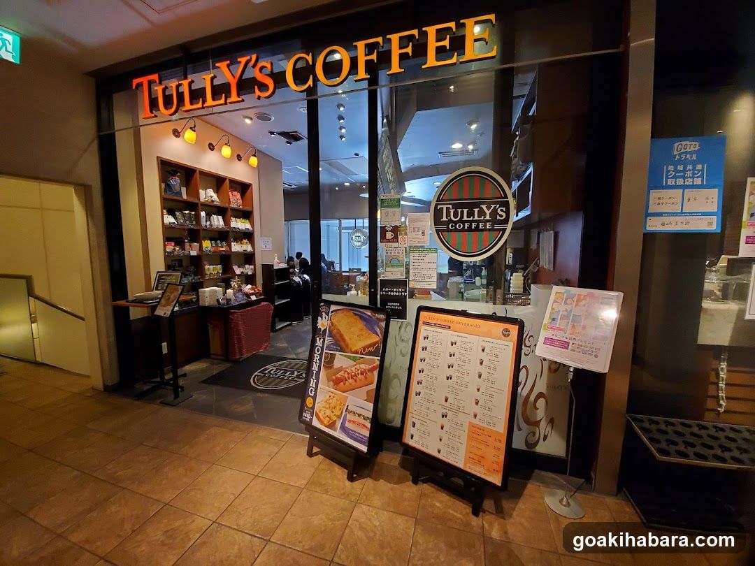 The image shows the exterior entrance of a Tully's Coffee shop, featuring illuminated signage above the glass doors and a circular logo on the window. Inside, shelves display coffee products, and staff are visible near the counter. Outside, there are menu boards with food items like sandwiches and pastries.