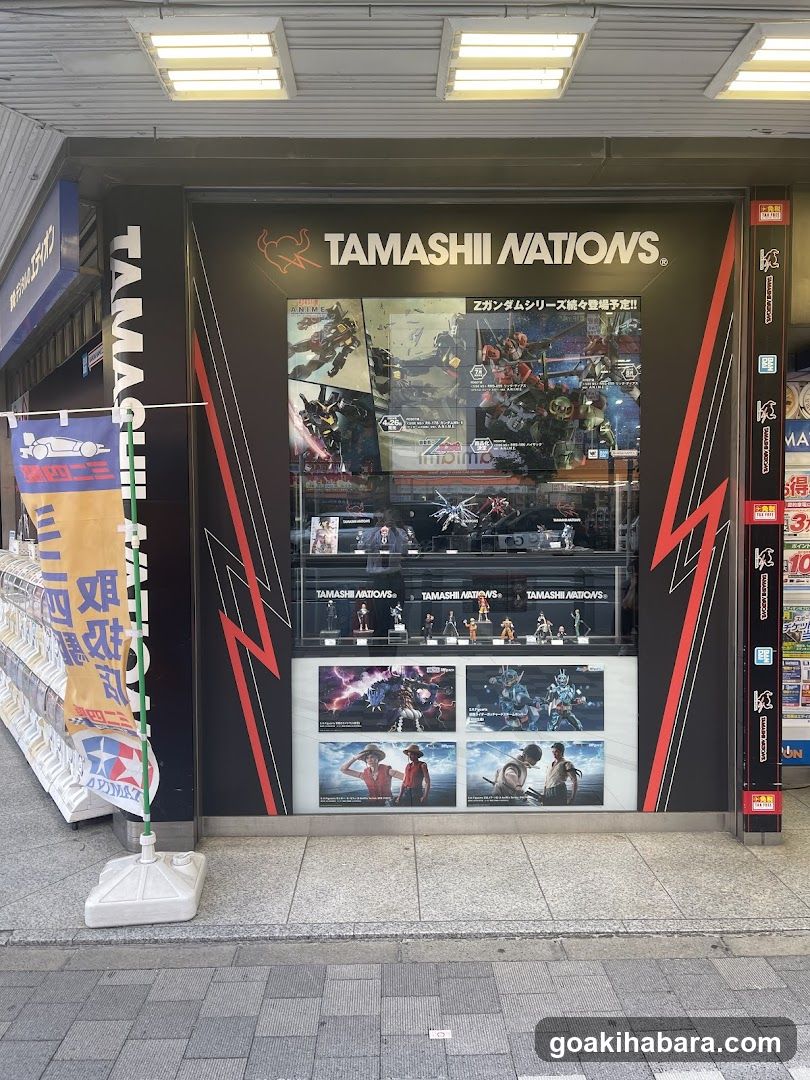 A storefront display featuring the TAMASHII NATIONS branding with a large window showcasing action figures and collectibles. The shop front includes vertical banners on either side, one reading 'TAMASHII NATIONS' in white letters against black background. Inside the display case are various figurines arranged neatly under bright lighting. Posters at the bottom highlight specific product lines or events.