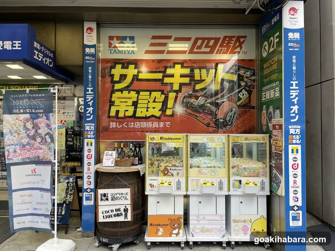 The image shows the storefront of a Tamiya shop, featuring a large banner advertising 'Mini-4WD' and 'Servo Kit'. There are three claw machines lined up in front of the store, each filled with colorful prizes. A barrel labeled 'Coco de Liquors' is placed near the entrance. The shop has various signs and advertisements, including one for 'Edion' and another promoting 'Watch Square'.