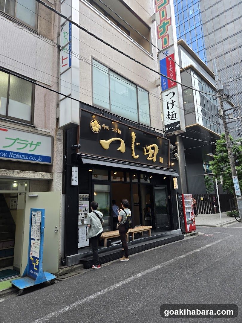 A street-level view of a ramen shop with black signage featuring gold Japanese characters. Two customers stand at the entrance, one holding a menu or pamphlet. A blue promotional stand is visible to the left, and a red vending machine stands near the sidewalk.