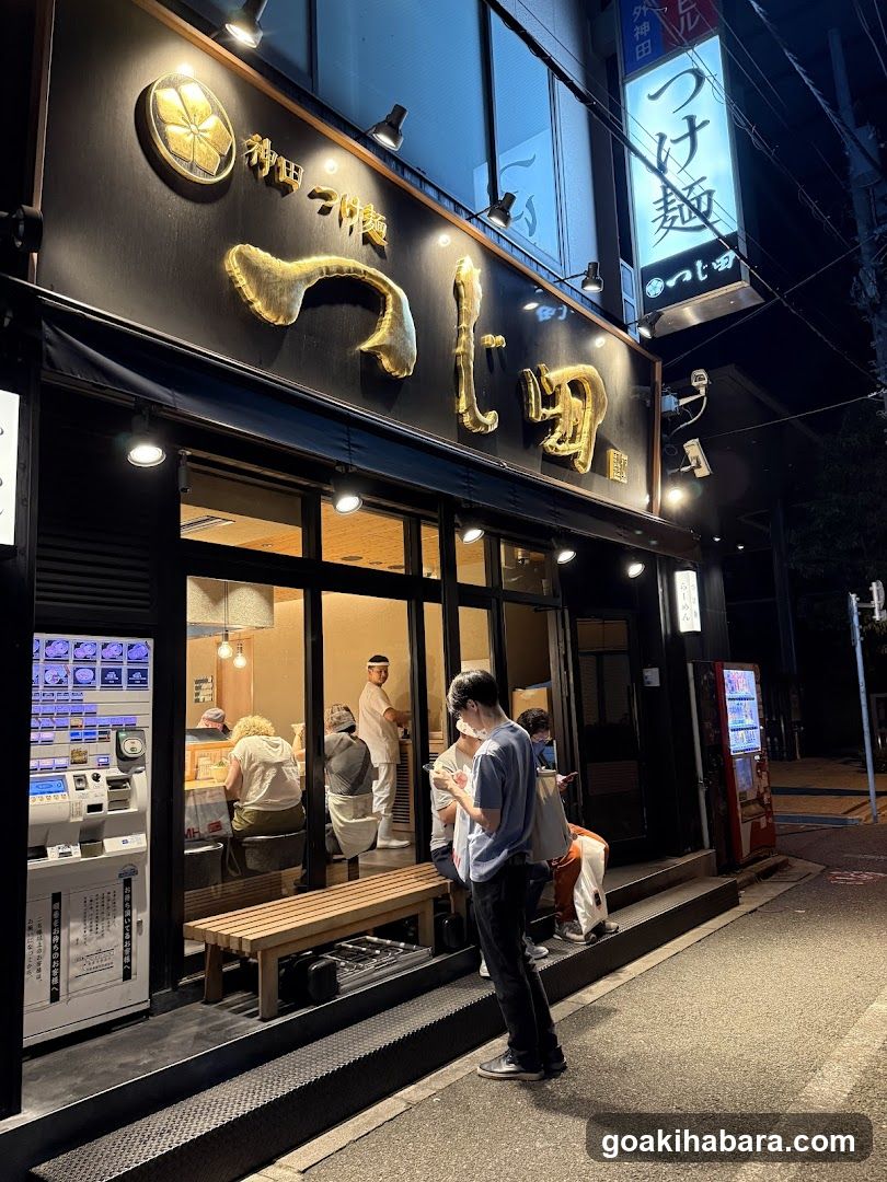A brightly lit shop front at night with large glass windows and doors, customers standing outside waiting or ordering, staff inside wearing chef hats, illuminated signage in Japanese characters above the entrance, vending machines on either side of the sidewalk.