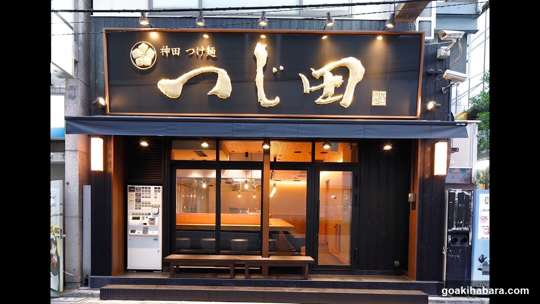 A modern shop front with a black facade and illuminated signage displaying 'つじ田' (Tsumi) in large gold characters. The entrance features glass doors revealing an interior with warm lighting, wooden benches outside, and a vending machine to the left.