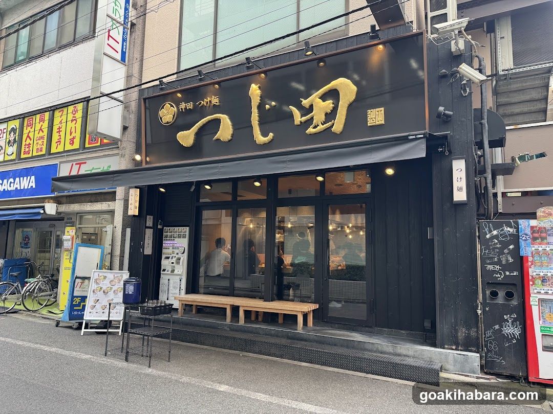 A street-level view of a Japanese restaurant named 'Tsujino' (つじ野) with prominent gold lettering on a black facade. The entrance features glass doors and windows, revealing interior seating. A wooden bench sits outside the entrance. To the left, there is a menu board displaying food items and prices. Adjacent buildings display various signs and advertisements.