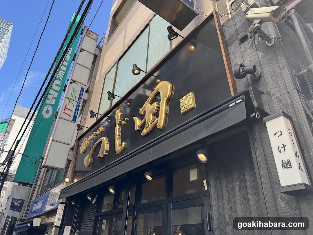 A street-level view of a ramen shop with large golden Japanese characters on the facade, illuminated spotlights, and vertical signage reading 'tsuke men' (dipping noodles). Adjacent buildings display signs including 'IT-club' and green banners.