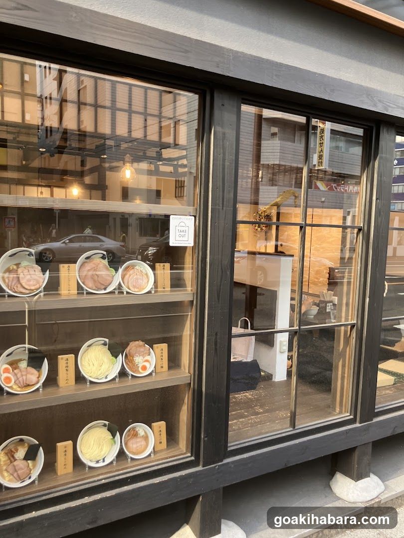 A shop front with large windows displaying ramen bowls on shelves inside, visible through the glass. A sign reading 'TAKE OUT' is affixed to one window pane. The interior shows wooden flooring and modern lighting fixtures. Reflections of surrounding buildings and vehicles are visible in the glass.