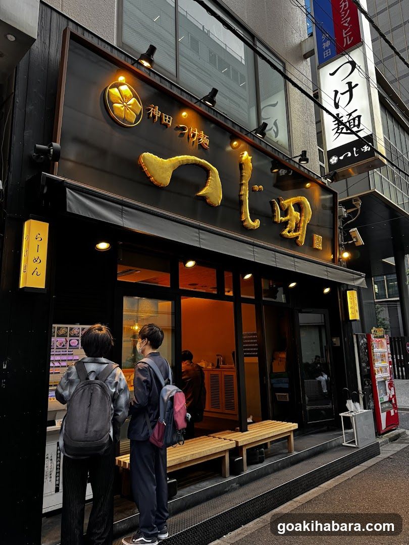 A street-level view of a ramen shop with black exterior and gold lettering. Two customers stand outside near the entrance, one wearing a backpack. A wooden bench sits at the doorway. Vertical signs display Japanese characters including 'ramen' and 'noodles'. Interior lighting is warm orange.