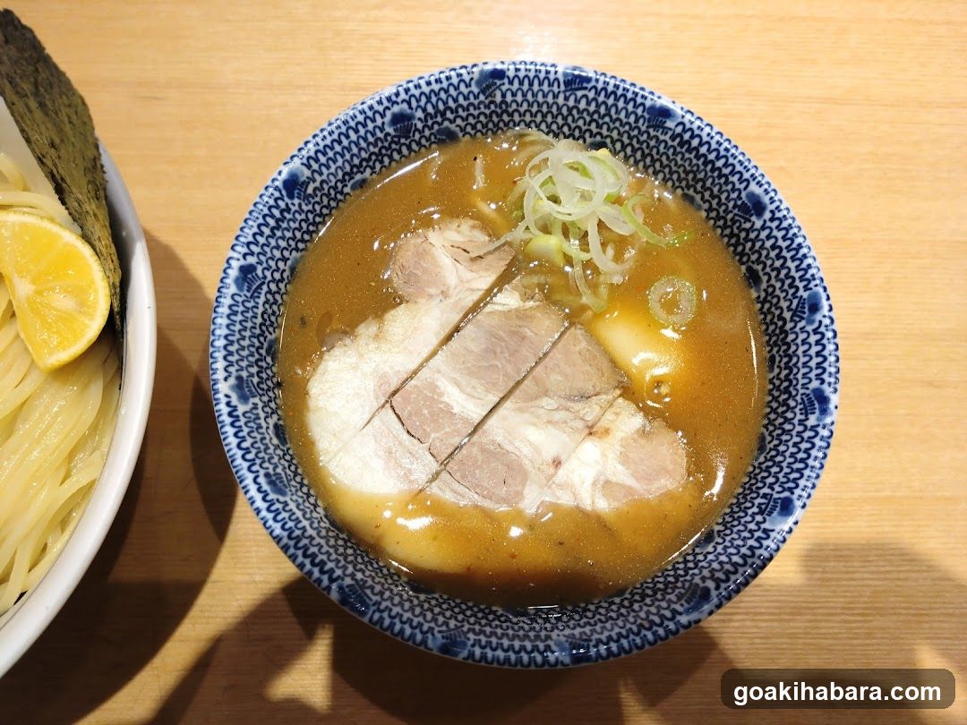 A bowl of ramen featuring sliced pork, green onions, and a rich broth in a blue patterned bowl, placed on a wooden table next to a side dish of noodles and lemon slices.