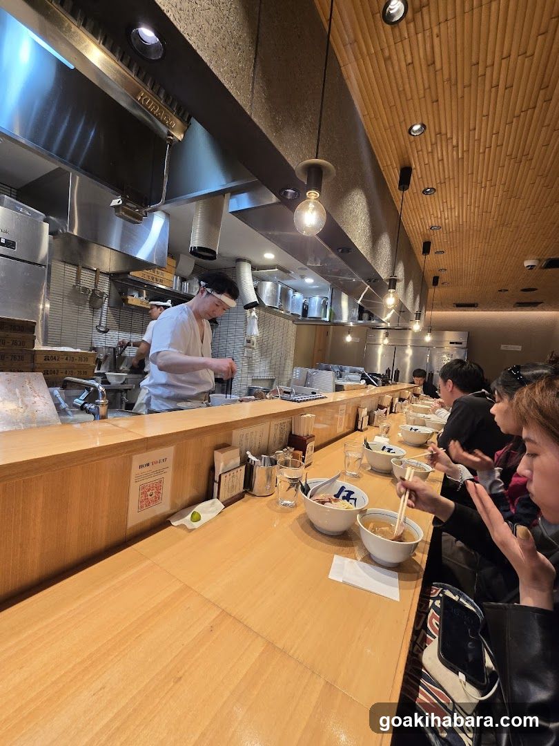 A cozy ramen bar with a long wooden counter where customers sit facing the kitchen. Chefs in white uniforms prepare food behind the counter, while diners enjoy bowls of ramen. The ceiling features bamboo-style paneling and hanging pendant lights. A QR code sign is visible on the counter.