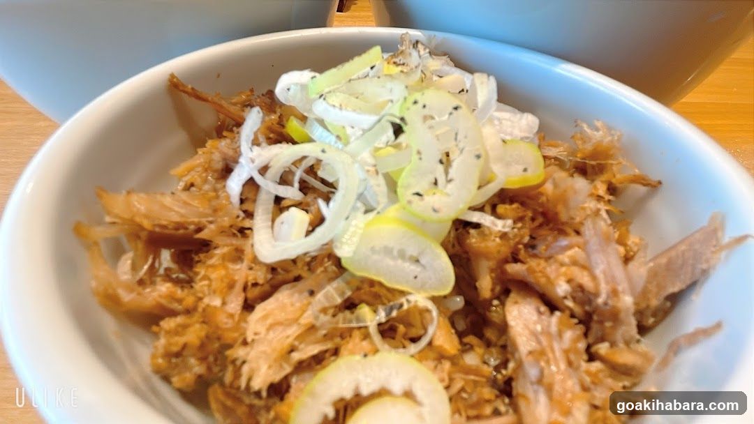 A close-up shot of a white bowl containing shredded browned meat, possibly pork or chicken, topped with sliced green onions and lemon slices.