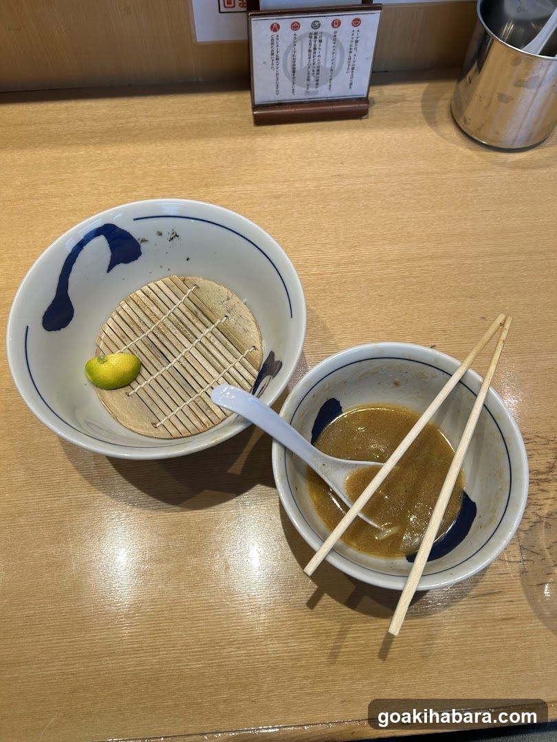 A close-up of a wooden table featuring two bowls. The larger bowl contains a small bamboo mat and a piece of lime, while the smaller bowl holds a brown broth with chopsticks resting on top. In the background, there is a menu card with Japanese text.