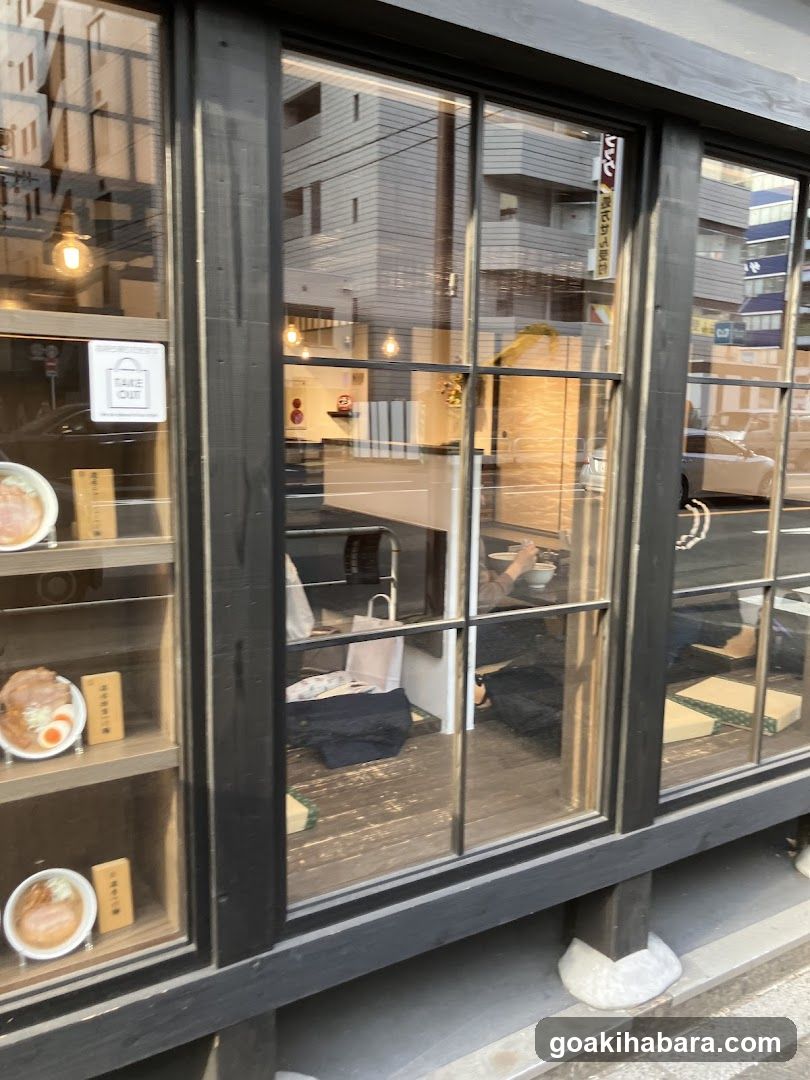 A photo of a shop's exterior entrance, showing large glass windows with dark frames. Inside, warm lighting illuminates the interior where customers are seated at tables. A 'TAKE OUT' sign is visible on the window. Reflections of surrounding buildings and street can be seen in the glass.