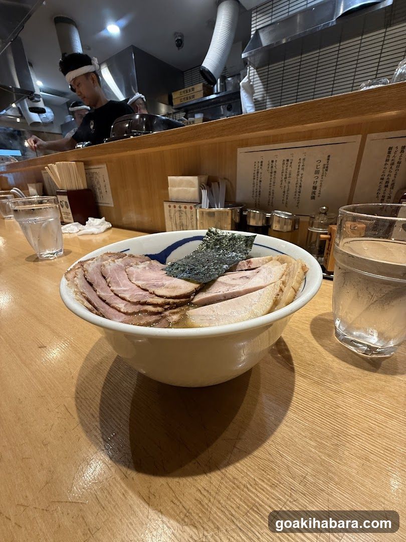 A bowl of ramen with sliced pork, seaweed, and broth sits on a wooden counter in a Japanese restaurant. A staff member is visible behind the counter preparing food.