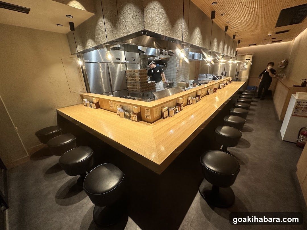 The image shows the interior of a restaurant featuring a long wooden bar counter lined with black stools. Behind the counter, stainless steel kitchen equipment and stacked boxes are visible. A chef in uniform is working near the center. To the right, another staff member stands near a white refrigerator. The ceiling has textured lighting fixtures casting warm illumination over the space.