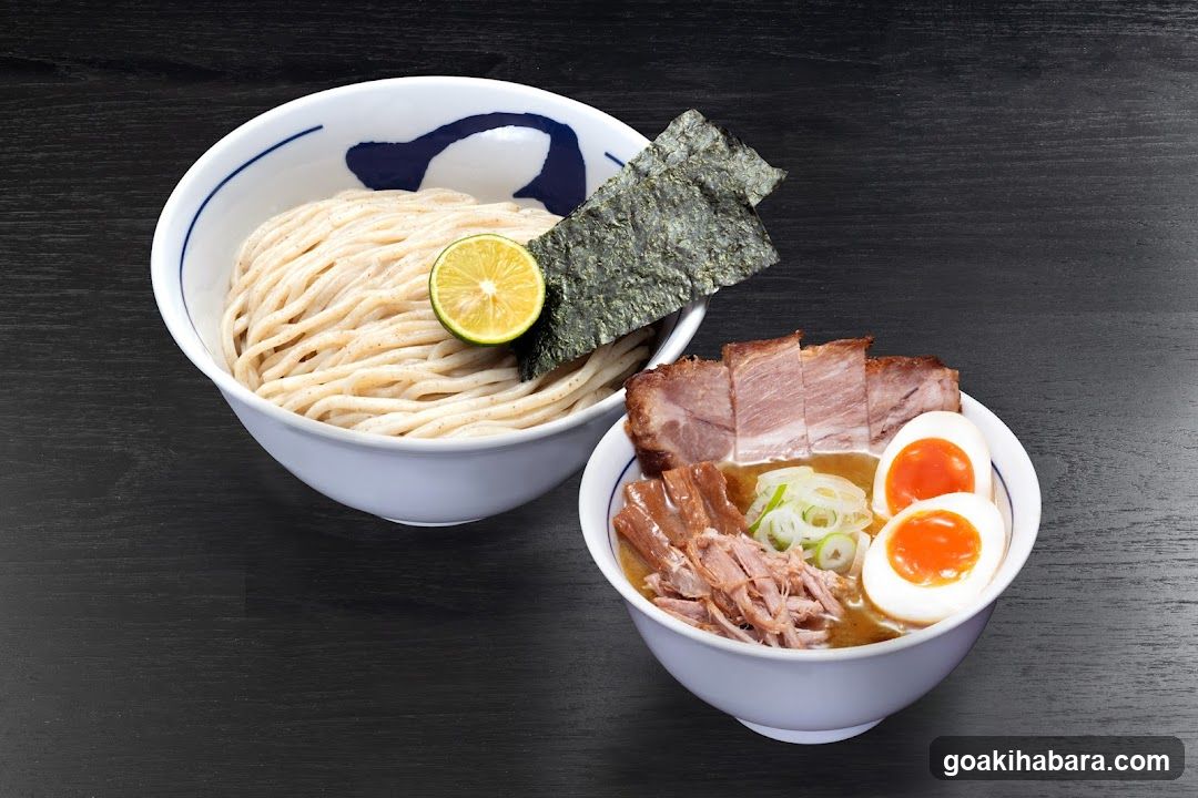 Two bowls of ramen on a dark wooden surface. The larger bowl contains noodles, a lime wedge, and nori sheets. The smaller bowl features broth, sliced pork, green onions, and halved soft-boiled eggs.