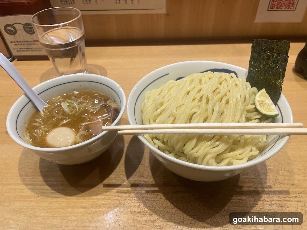 A table setting featuring a bowl of ramen with broth, green onions, and a soft-boiled egg, alongside a bowl of plain noodles garnished with seaweed and lime wedge. A glass of water is placed nearby.