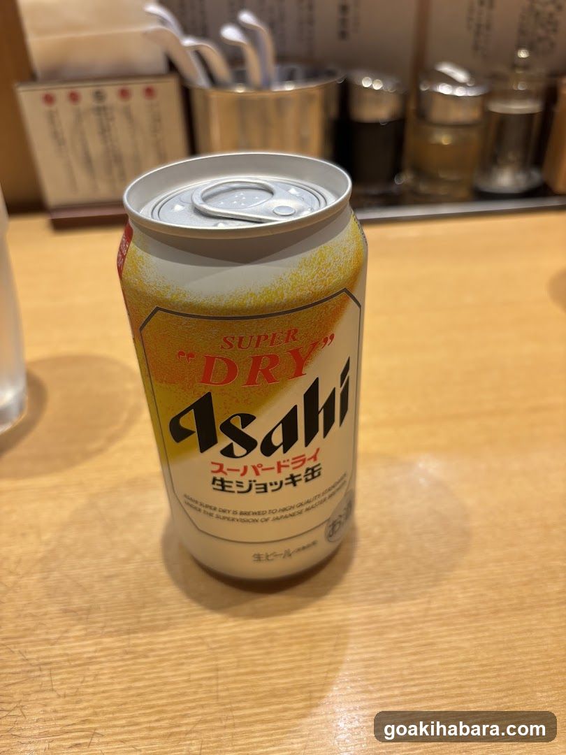 A close-up photo of an Asahi Super Dry beer can sitting on a wooden table in what appears to be a restaurant or bar setting. The background includes condiment shakers and cutlery holders, suggesting a dining environment.