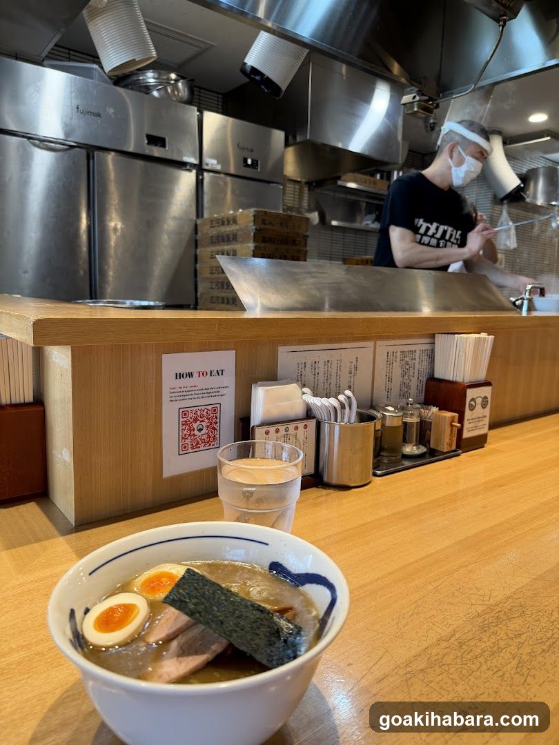 A bowl of ramen with soft-boiled egg and seaweed sits on a wooden counter in a Japanese restaurant. Behind the counter, stainless steel kitchen equipment is visible, along with a chef wearing a mask and headband preparing food. A QR code sign labeled 'HOW TO EAT' is posted on the counter.