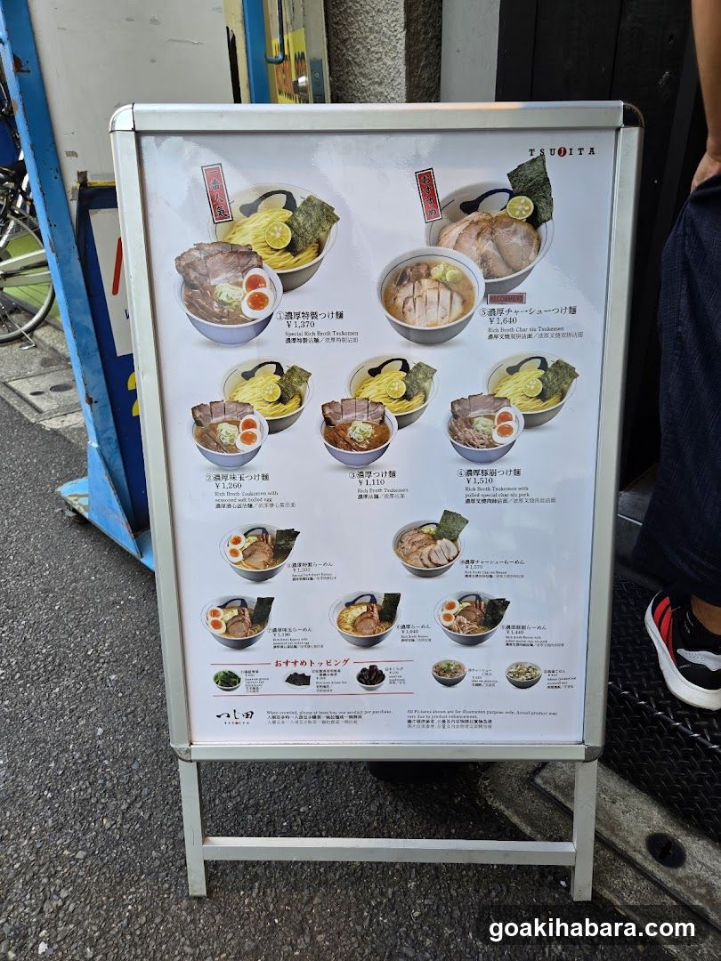 A standing menu board outside a ramen shop displaying various noodle dishes with images, names in Japanese and English, prices, and recommended toppings.