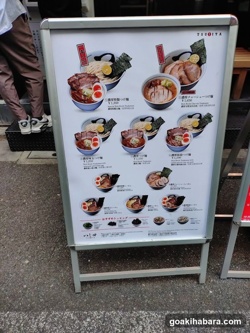 A-frame menu board displaying various ramen dishes with prices in yen, featuring images of bowls and toppings like pork slices, eggs, and seaweed.