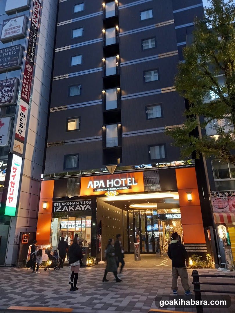 Evening exterior shot of the APA Hotel entrance featuring illuminated signage and a visible izakaya storefront with people walking by.