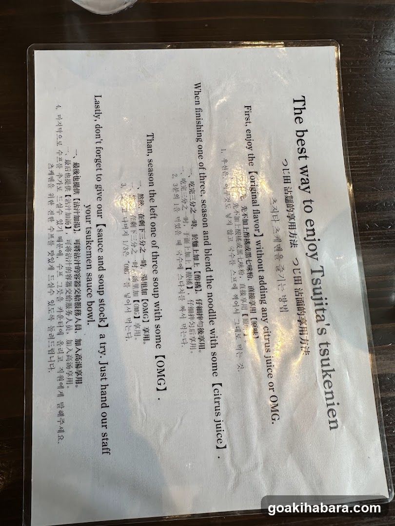 A printed menu card on a dark wooden table, featuring instructions and recipes for preparing Tsukune (Japanese meatballs). The card includes English and Korean text with step-by-step cooking directions.