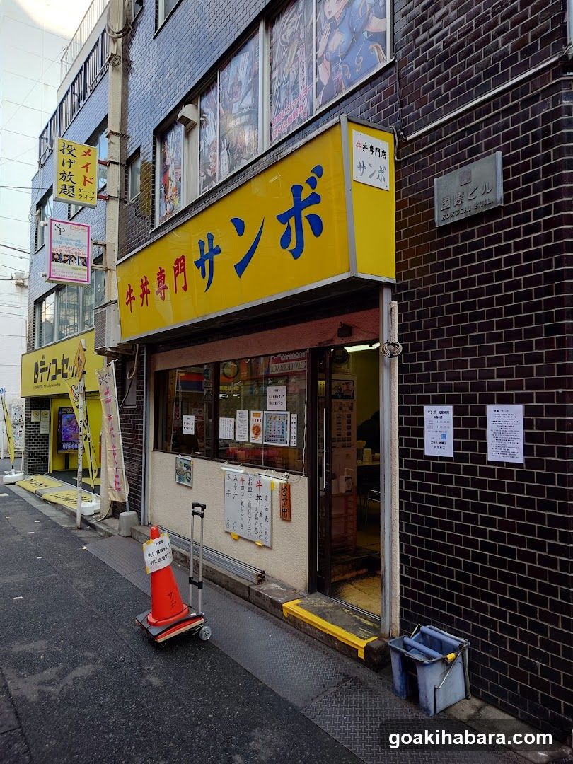 A street-level view of a Japanese shop with a yellow sign reading 'Nikkei Sanbo' and smaller signs indicating 'Wagyu Special'. The entrance is visible, with posters on the wall and a menu board outside.