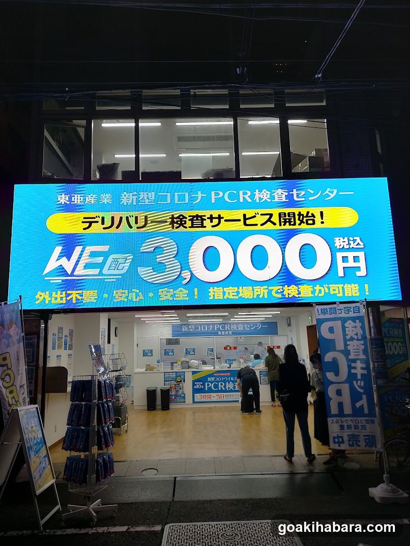 Night exterior shot of a medical facility storefront with large LED signage advertising delivery-based PCR testing services starting at ¥3,000 (tax included). The sign features Japanese and English text emphasizing safety and convenience without leaving home. Inside, customers are seen waiting near reception counters under bright fluorescent lighting. Promotional banners flank the entrance.