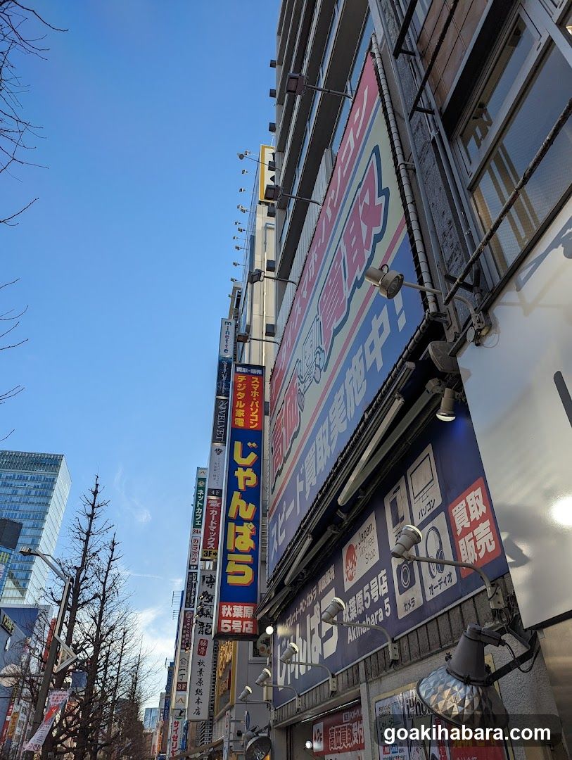 A low-angle shot of a busy city street lined with multi-story buildings featuring numerous vertical and horizontal signs in Japanese characters. Prominent signage includes 'ジャンボ' (Jumbo) and '買取実施中!' (Buying services active!). The scene captures the dense commercial atmosphere typical of Tokyo's shopping districts, with clear blue skies above bare tree branches.