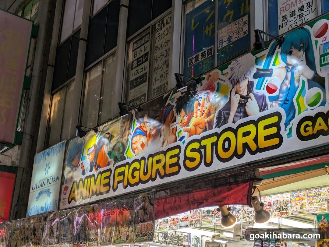 Night shot of a storefront with large anime character posters above the entrance, including Naruto and Hatsune Miku. Signage reads 'ANIME FIGURE STORE' in bold yellow letters. Below, shelves display packaged figures. Vertical signs on building show store hours and product categories like ROM models and figure parts.