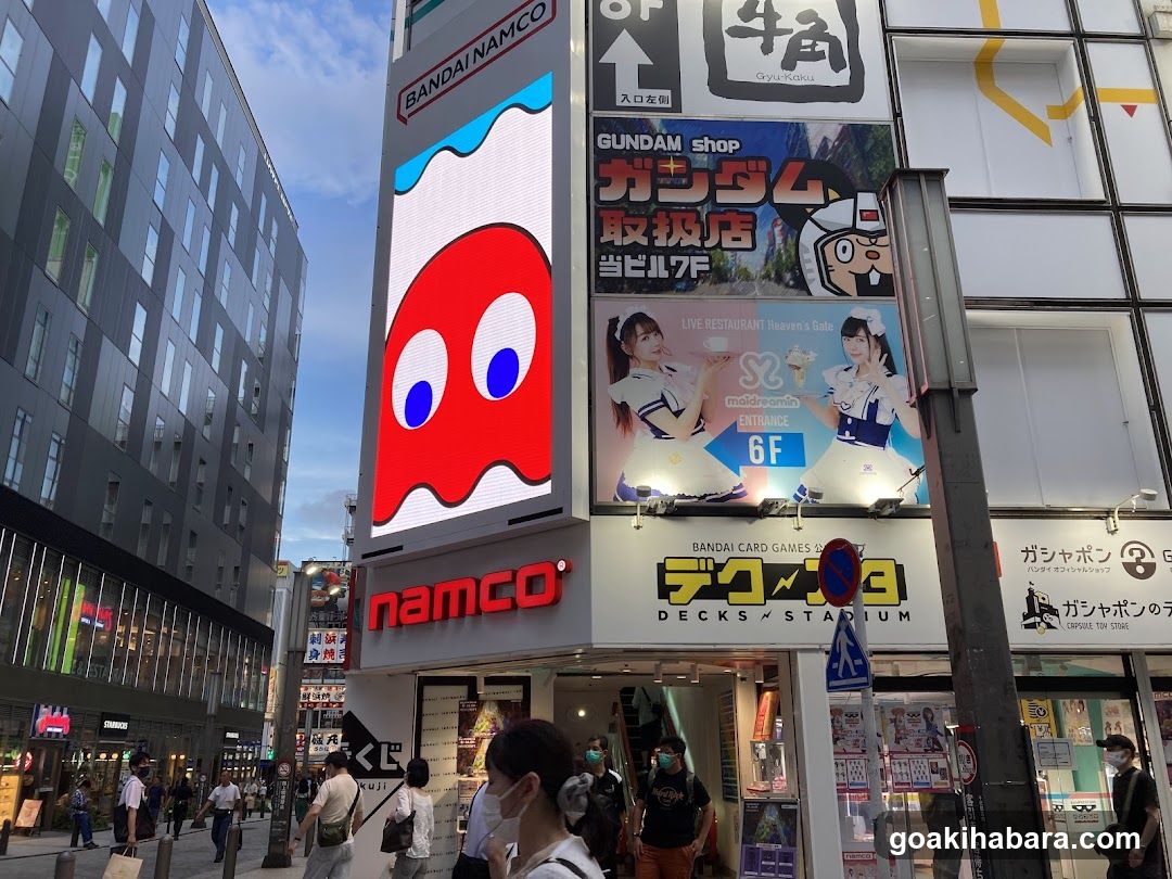 Street-level view of a multi-story building in Japan featuring prominent signage for Namco and Bandai Namco. The facade includes illuminated signs for 'Gundam Shop', 'Live Restaurant Heaven's Gate', and 'Bandai Card Games'. People are walking on the sidewalk below, with modern buildings visible in the background under a partly cloudy sky.