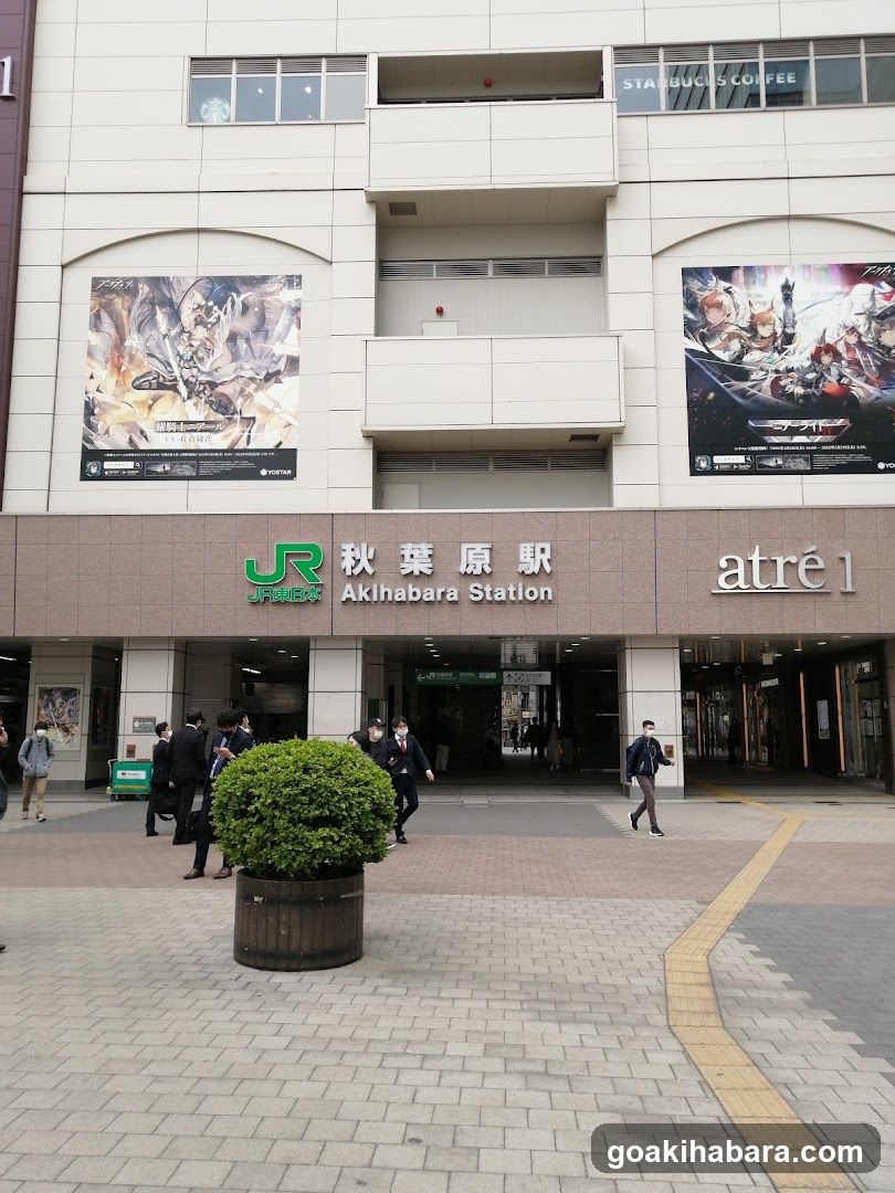 The image shows the exterior entrance of Akihabara Station, featuring a modern building facade with large posters advertising anime games. The JR logo and station name are prominently displayed above the entrance, along with signage for atré1.