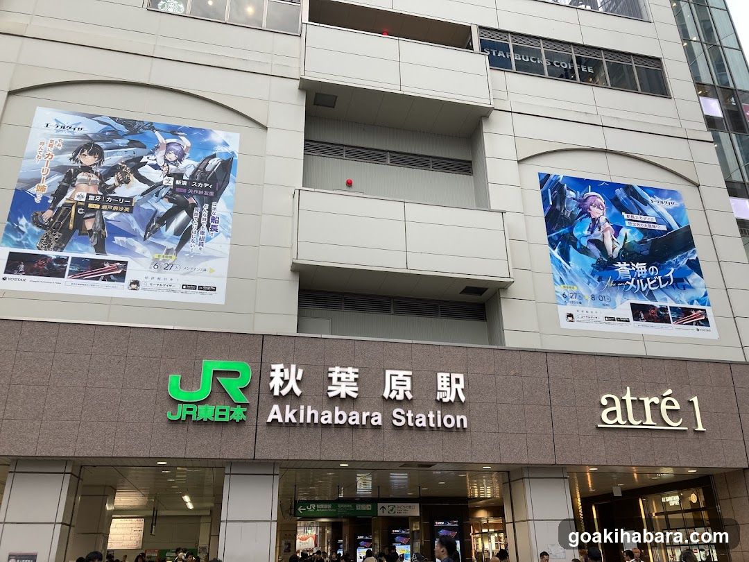 Photo of the physical exterior entrance and storefront facade of Akihabara Station in Japan. Visible signage includes JR East logo, '秋葉原駅' (Akihabara Station), 'Akihabara Station', and 'atré1'. Large anime-themed posters are displayed on the building walls above the entrance.