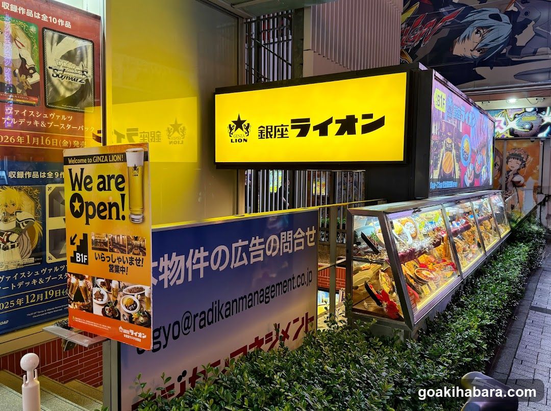 Night shot of Ginza Lion storefront with illuminated yellow sign, glass display cases filled with food items, and multiple posters advertising events and menu specials.