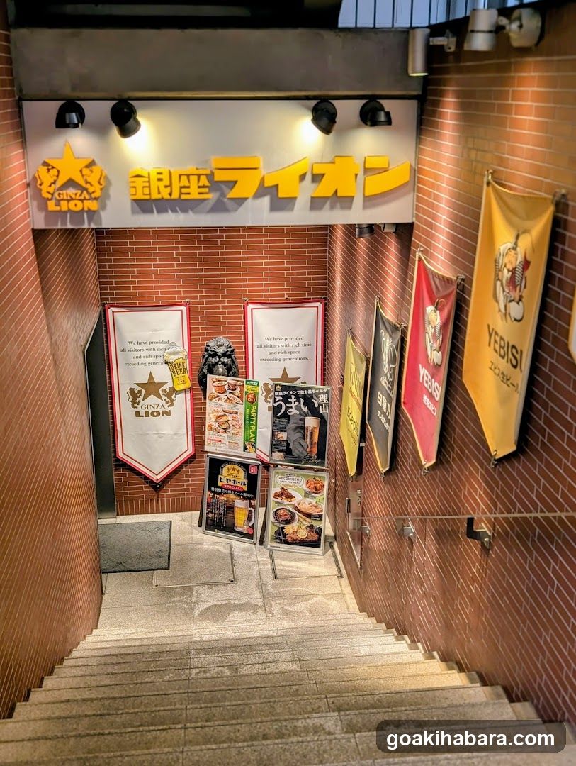 A downward view of a brick-walled stairway entrance to the Ginza Lion restaurant, featuring illuminated signage with Japanese and English text, banners on the right wall, and promotional posters near the bottom.