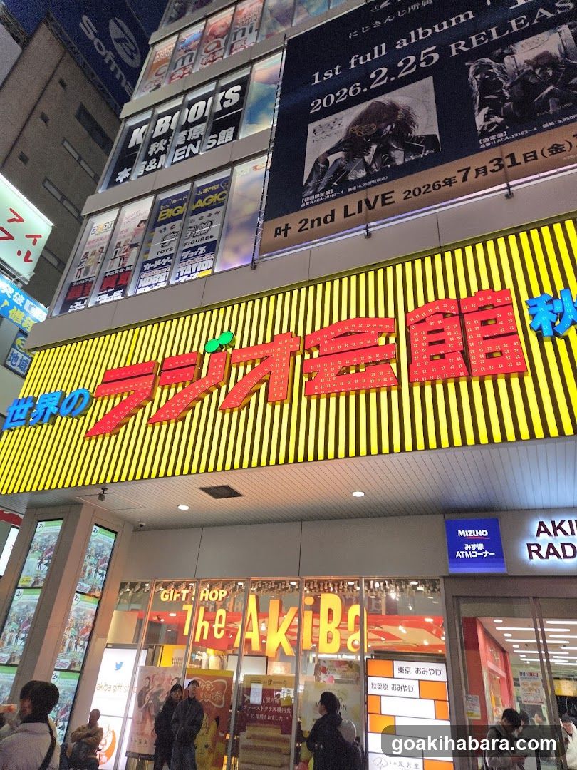 A brightly lit storefront with yellow vertical stripes and red neon signage reading 'ラジオ会館' (Radio Kaikan). Above, banners advertise music releases and live events. The entrance features glass doors with promotional posters inside. People are visible near the entrance.