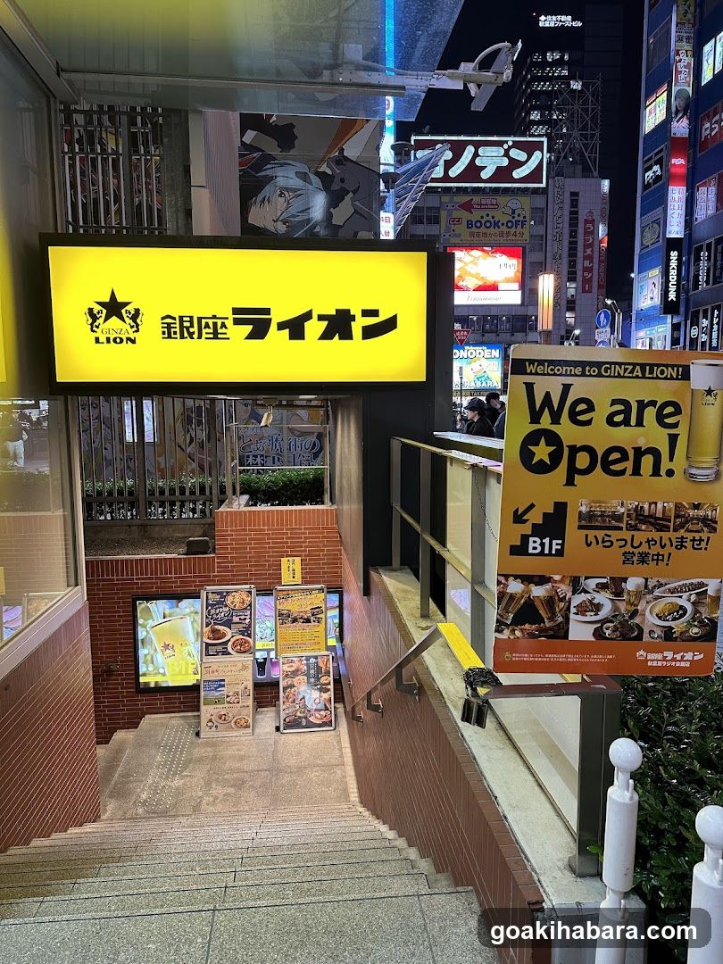 Night view of the Ginza Lion restaurant entrance, featuring a bright yellow sign with '銀座ライオン' and a star logo. A staircase leads down to the entrance, flanked by illuminated menu boards and promotional posters. The surrounding area is bustling with neon signs and city lights.